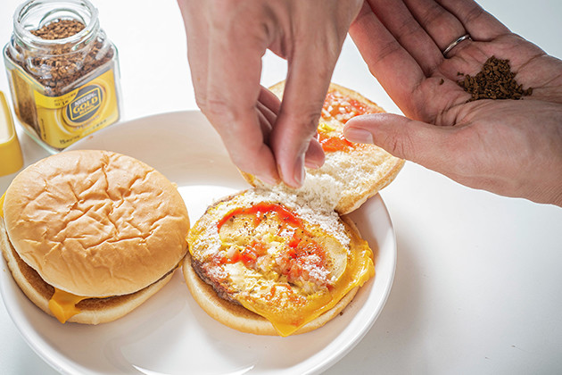 （１）かける！　チーズバーガーを開き、指先でネスカフェゴールドブレンドをつまみ、砕きながらビーフパティ全体に軽く振りかける。バンズで再びビーフパティを挟んだら完成だ